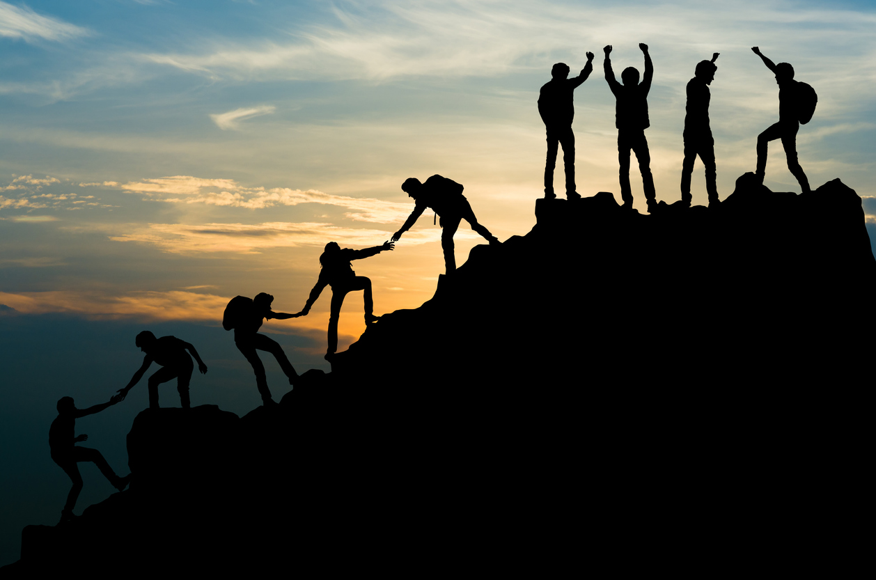 Silhouetted group of hikers helping one another climb a steep mountain at sunrise, symbolizing trust, cooperation, and paying attention to others’ actions.