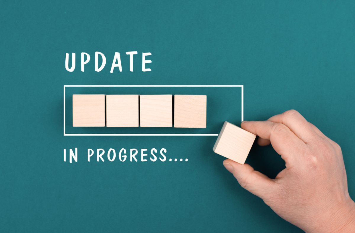 Top-down view of a hand placing wooden blocks inside a white progress bar on a teal background with the words ‘UPDATE’ above and ‘IN PROGRESS….’ below.