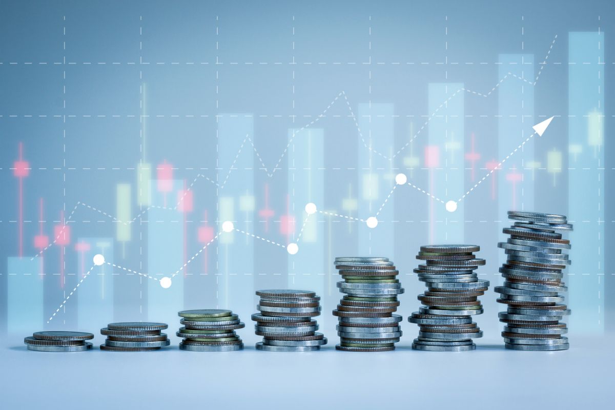 Stacks of coins increasing in height from left to right in front of a faint stock chart with candlesticks and an upward-trending line, symbolizing investment growth and rising returns.