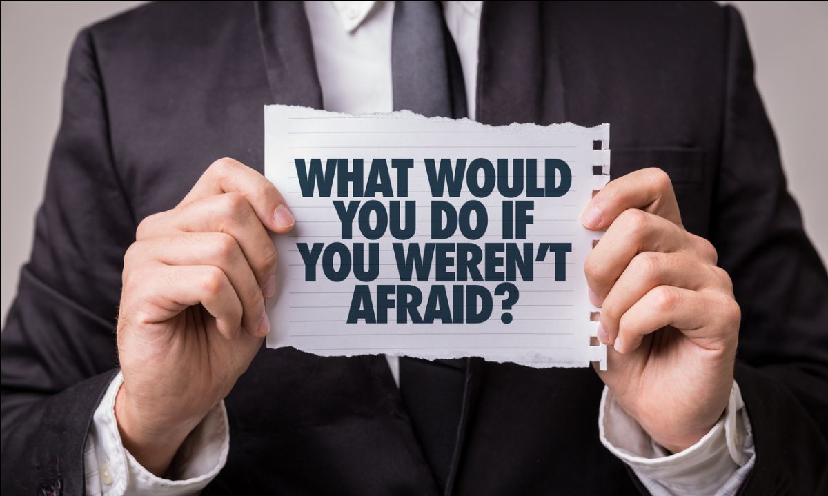 Person in a suit holding a torn sheet of paper that reads ‘WHAT WOULD YOU DO IF YOU WEREN’T AFRAID?’ in bold capital letters