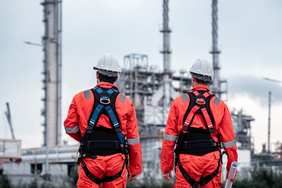 “Two industrial workers in orange safety gear and white hard hats standing with their backs to the camera, looking at large refinery or petrochemical plant structures in the distance.”