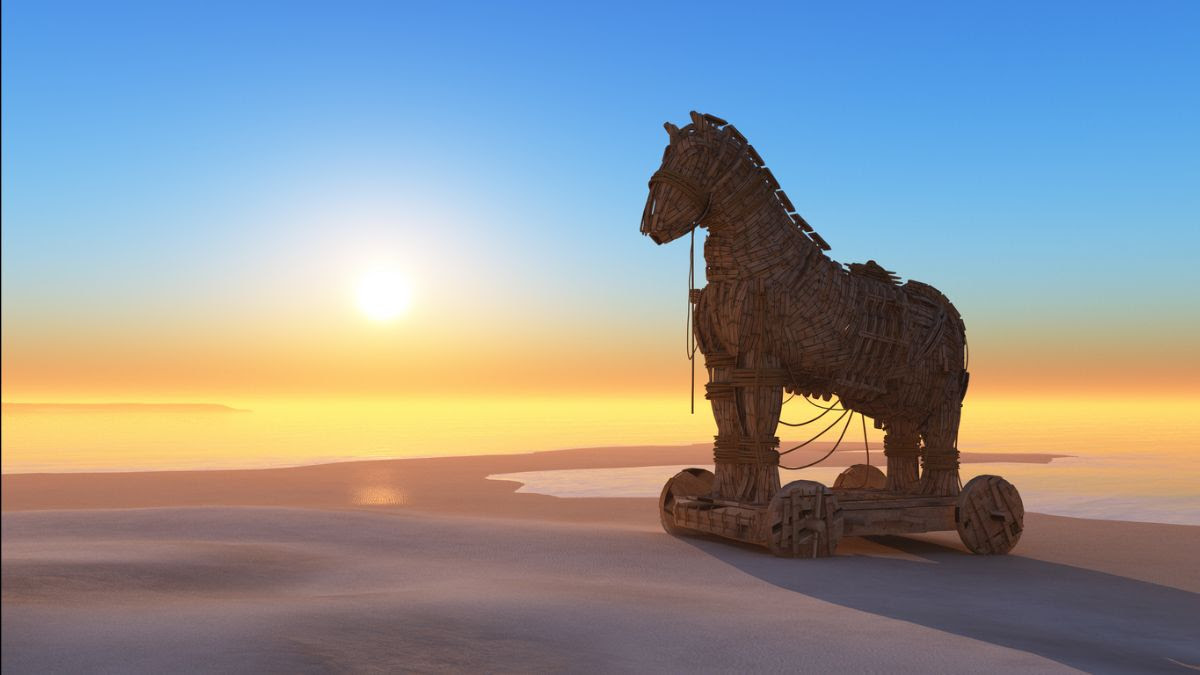 A wooden Trojan horse statue on wheels standing on a sandy shore at sunrise, with the sun low over a calm sea and a clear blue-to-gold sky