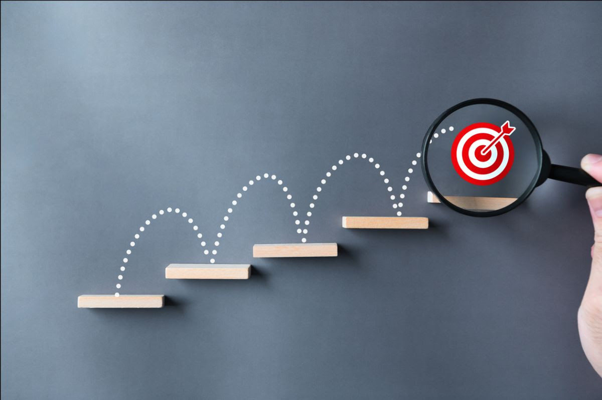 An illustration of wooden steps with a dotted bouncing path leading to a red bullseye target under a magnifying glass, symbolizing progress toward a goal.